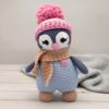 A cute crocheted penguin wearing a pink hat with a pom-pom, a beige scarf, and a small heart detail stands on a light wooden surface.