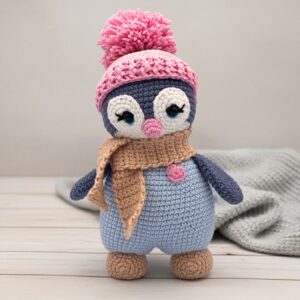 A cute crocheted penguin wearing a pink hat with a pom-pom, a beige scarf, and a small heart detail stands on a light wooden surface.