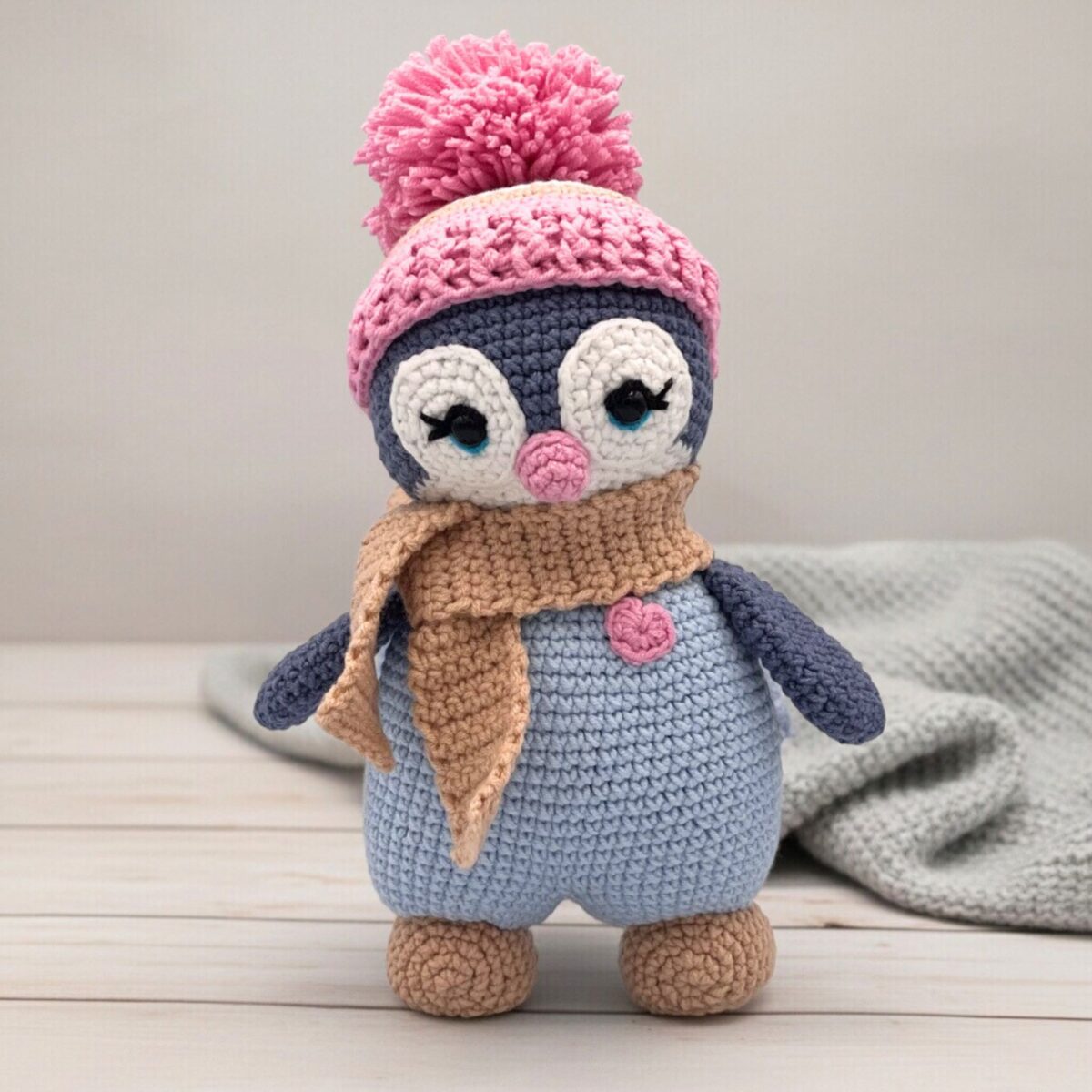 A cute crocheted penguin wearing a pink hat with a pom-pom, a beige scarf, and a small heart detail stands on a light wooden surface.