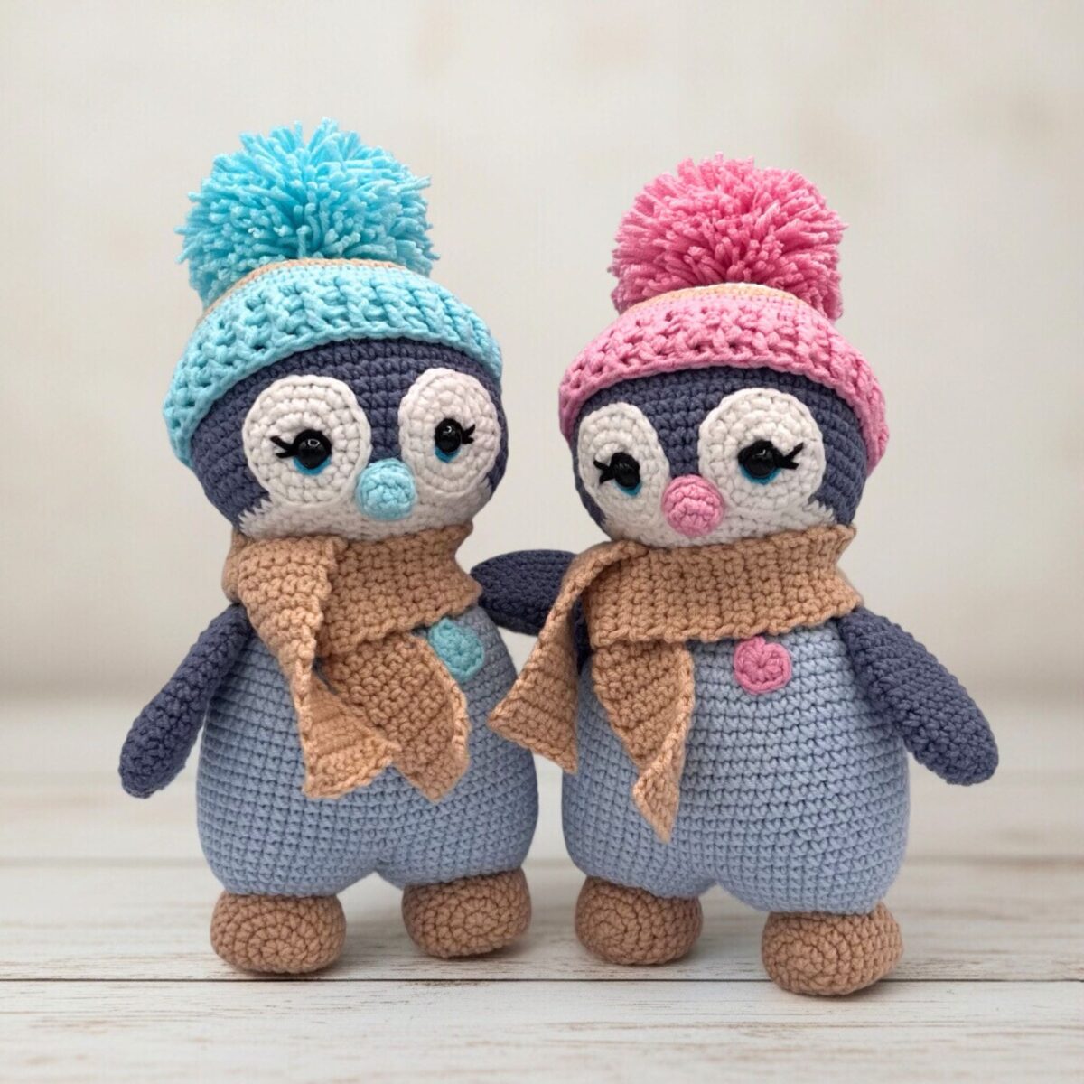 Two crocheted penguins wearing colorful hats with pompoms and matching scarves stand side by side on a wooden surface, conveying warmth and friendship.