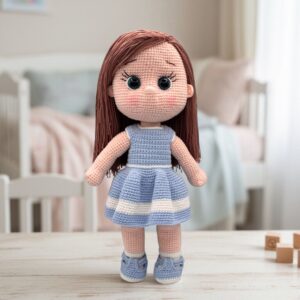 A crochet doll with brown hair, large black eyes, and a blue and white striped dress stands on a wooden table. Background shows a softly lit bedroom.