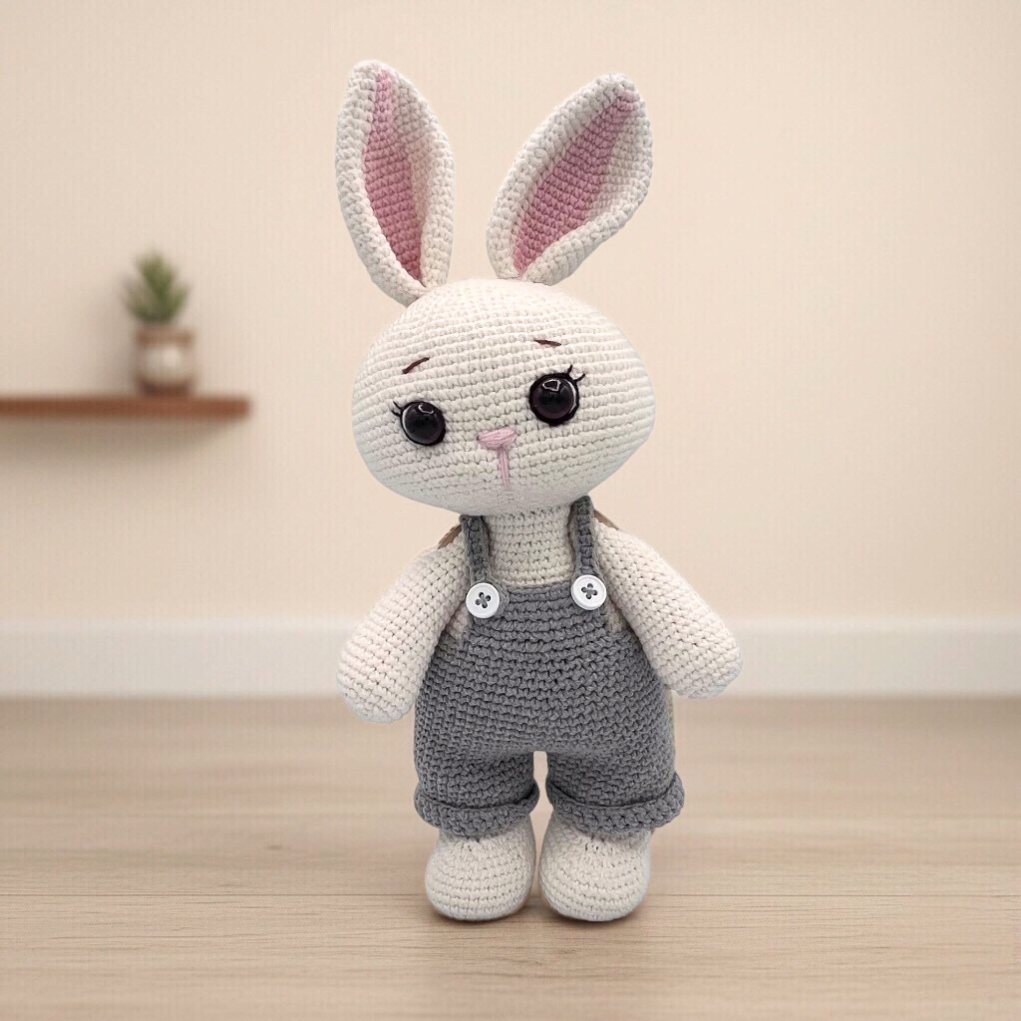 Crocheted white bunny with large dark brown eyes and pink ears, wearing gray overalls with buttons. Soft focus background hints at an Easter-themed setting.