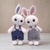Two crocheted bunny toys wearing overalls stand side by side. One wears navy blue and the other gray.