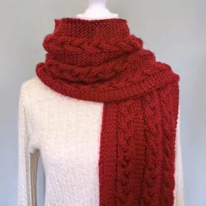 A vibrant red, thick knitted scarf with intricate cable patterns is draped over a beige sweater. The scene conveys cozy warmth and elegance.