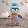 A crocheted penguin toy with blue and beige colors wears a beanie and scarf, standing on wooden floor. Soft decor with stars and cloud in background. Cozy and playful tone.