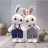 Two crocheted bunny toys wearing overalls stand side by side. One wears navy blue and the other gray. A basket with pastel eggs is in the background.