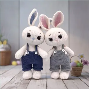 Two crocheted bunny toys wearing overalls stand side by side. One wears navy blue and the other gray. A basket with pastel eggs is in the background.