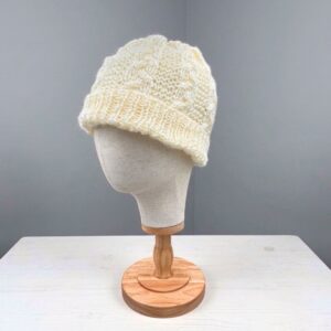 Knitted beanie with a folded ribbed brim and a vertical cable‑knit pattern, displayed on a beige mannequin head mounted on a wooden stand against a plain gray background
