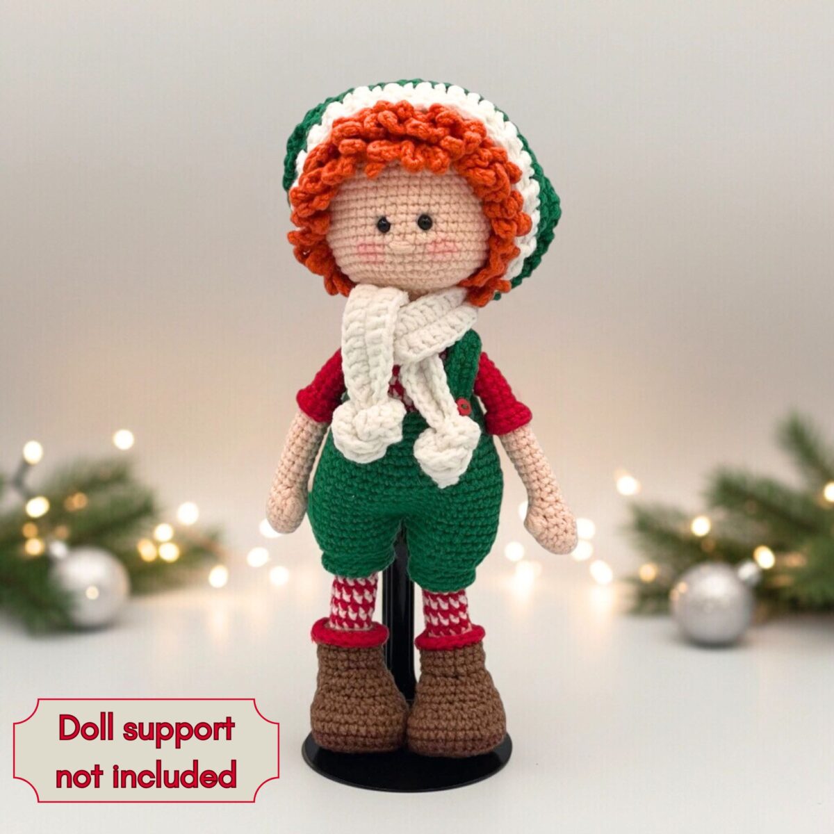 Crochet doll with curly orange hair dressed in festive green outfit and white scarf. Soft lighting and greenery in background evoke holiday spirit.