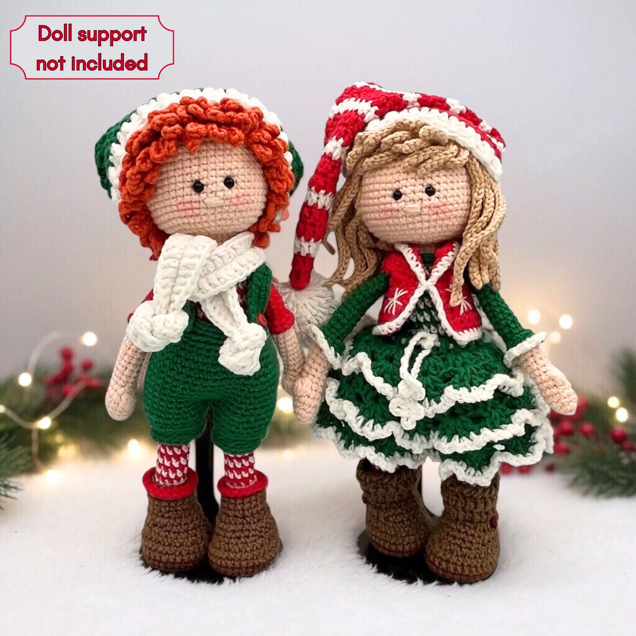 Two crocheted dolls in festive attire stand hand in hand. The left doll has curly orange hair and a white scarf. The right doll wears a striped hat and green dress. Festive lights and pine branches are blurred in the background.