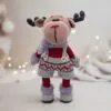 A crocheted reindeer toy stands upright, wearing festive burgundy and gray overalls with patterned trim. Soft, cozy textures create a warm holiday vibe.