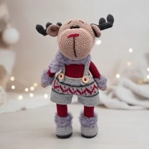 A crocheted reindeer toy stands upright, wearing festive burgundy and gray overalls with patterned trim. Soft, cozy textures create a warm holiday vibe.