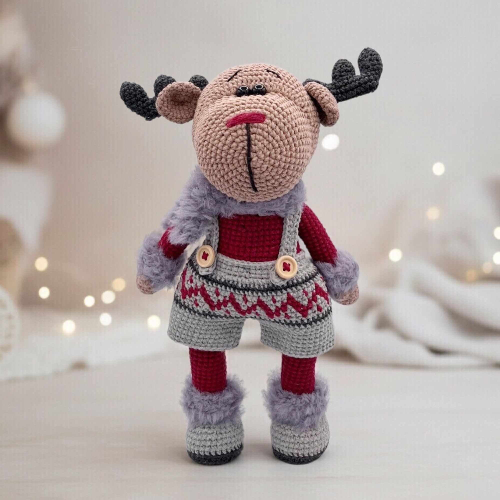 A crocheted reindeer toy stands upright, wearing festive burgundy and gray overalls with patterned trim. Soft, cozy textures create a warm holiday vibe.