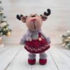 A crocheted reindeer doll dressed in a burgundy dress and grey scarf stands on a wooden surface. Christmas trees with lights are blurred in the background.