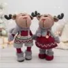 Two crocheted reindeer dolls, dressed in festive red and gray outfits with antlers, stand on a wooden surface against a cozy, holiday-lit background.