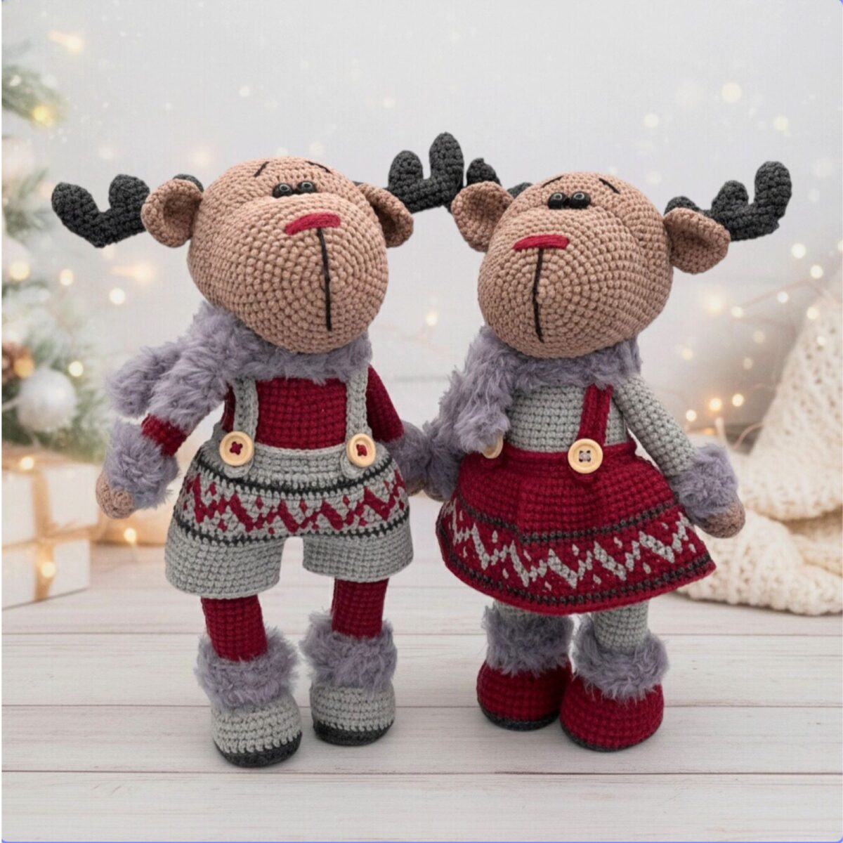Two crocheted reindeer dolls, dressed in festive red and gray outfits with antlers, stand on a wooden surface against a cozy, holiday-lit background.