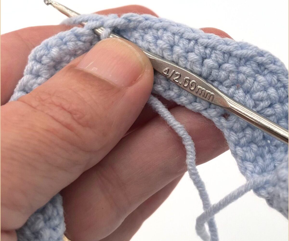 Close-up of Ruth's hand crocheting with a blue-gray yarn, showing the crochet hook size