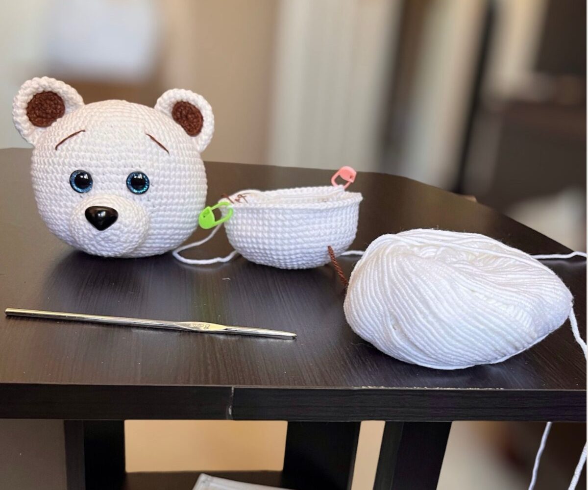 An amigurumi bear head in progress on Ruth's work table, with white yarn and a crochet hook