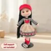 Crocheted doll with long black hair, wearing a red hat and heart-patterned outfit, stands on a black support.