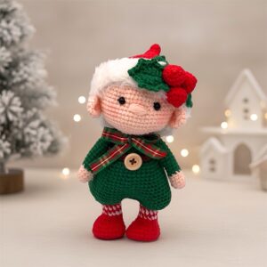 Crocheted doll in a green outfit with red boots and a white hat, decorated with holly. Stands in front of a snow-covered tree and white buildings. Holiday vibe.
