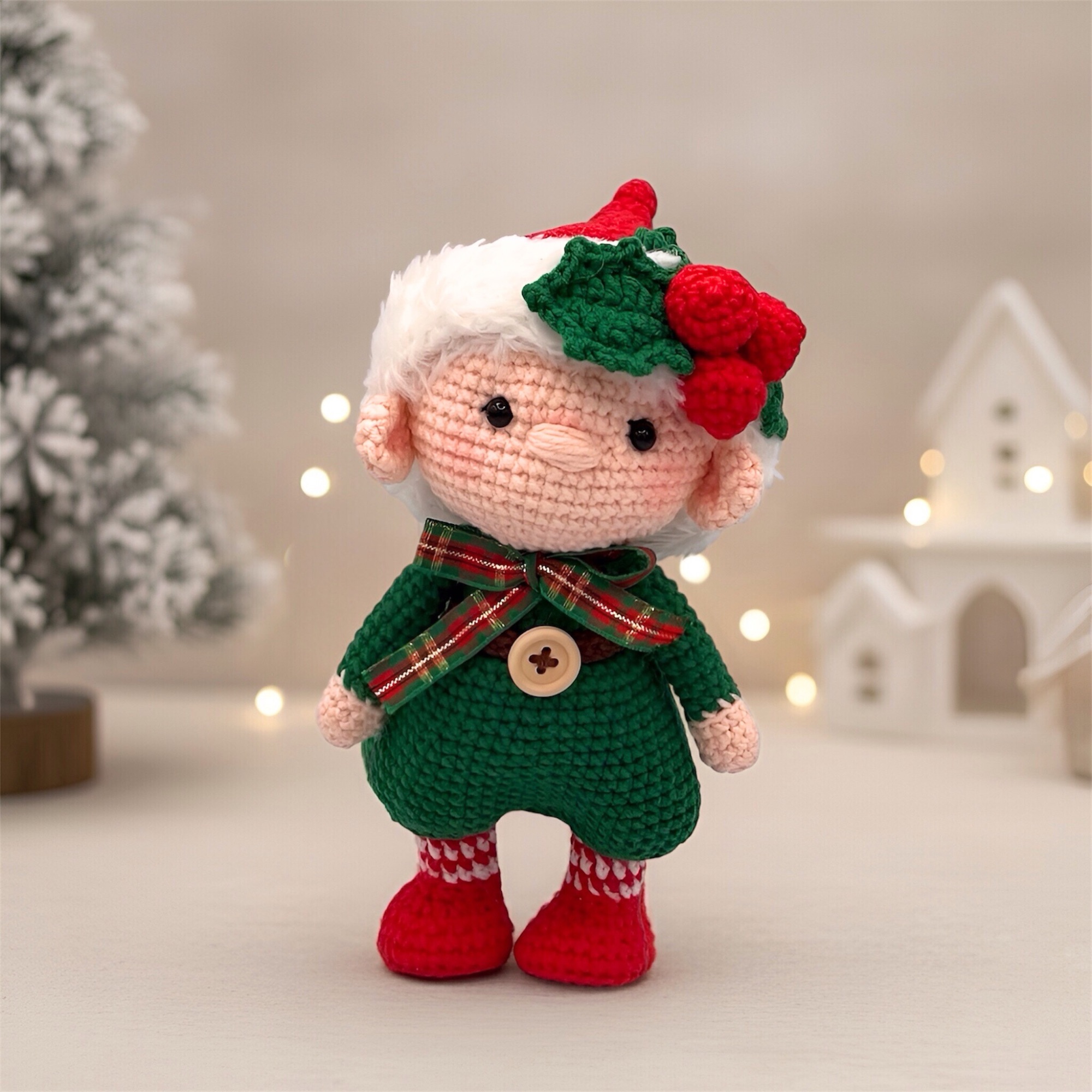 Crocheted doll in a green outfit with red boots and a white hat, decorated with holly. Stands in front of a snow-covered tree and white buildings. Holiday vibe.