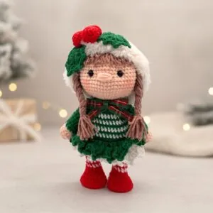Crochet doll in festive attire with a green dress, red boots, and holly on a hat. Soft lighting and a blurred background create a cozy feel.