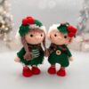 Two crocheted dolls in festive green outfits with red berries and holly accents stand side by side, set against a blurred, light-filled background.