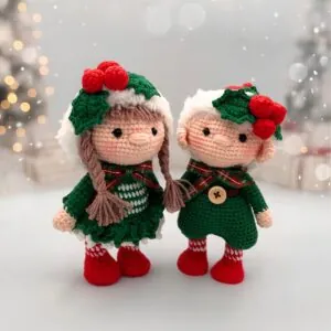 Two crocheted dolls in festive green outfits with red berries and holly accents stand side by side, set against a blurred, light-filled background.