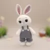 Crocheted white bunny with large dark brown eyes and pink ears, wearing gray overalls with buttons. Soft focus background hints at an Easter-themed setting.