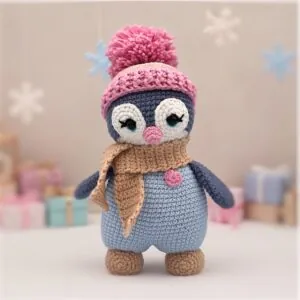 Cute crochet penguin plush in winter attire, featuring a pink beanie with pompom and a beige scarf. Soft pastels create a cozy, festive vibe.