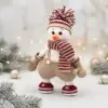 A crocheted snowman wearing a burgundy and white striped scarf and hat, with mitten-like hands and boots, stands amidst soft-focus twinkling lights and frosted pine branches, conveying a cozy and cheerful winter scene.