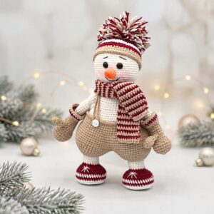A crocheted snowman wearing a burgundy and white striped scarf and hat, with mitten-like hands and boots, stands amidst soft-focus twinkling lights and frosted pine branches, conveying a cozy and cheerful winter scene.