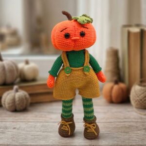 A handmade crochet doll with an orange pumpkin head, green shirt, yellow overalls with buttons, striped leggings, and brown boots on a wooden table.