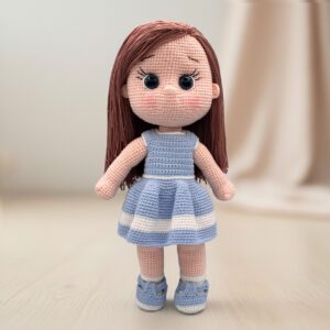 A crocheted doll with long brown hair and large black eyes stands on a light surface. She's wearing a blue and white striped dress and matching shoes.