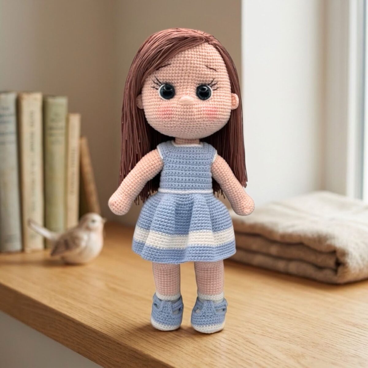 A crocheted doll with long brown hair and large black eyes stands on a light surface. She's wearing a blue and white striped dress and matching shoes.